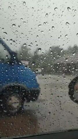 The video shows a car driving on a rainy day. The view is from inside the car, looking out through the windshield, which is covered in raindrops. There are other cars and buildings visible through the rain. The side mirror is also visible through the side window, also covered in raindrops. The wipers are on.