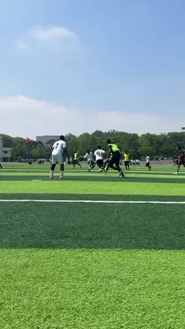 The video shows a group of men playing soccer on a green, synthetic turf field. Several men are wearing white shirts with black shorts. One man has a lime green vest over a black shirt. Players run, kick, and chase after a soccer ball. In the background are trees, buildings, and spectators. The weather is sunny.