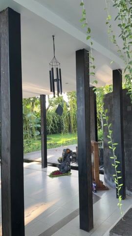The video shows a partially enclosed outdoor space with black wooden support beams. Hanging from the white ceiling is a black wind chime. A small black statue sits in the center of the frame, surrounded by greenery and a few flowers. Beyond the enclosed area is a lush green yard with trees and a wooden walkway. Vines hang from the side of the enclosure. The space appears to be well-lit with natural light. There are no people visible in the video.