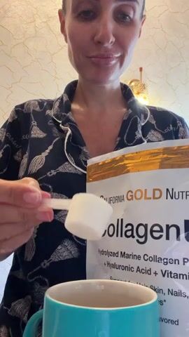 A woman in pajamas is shown from the chest up, pouring a white powder from a scoop into a teal mug. She is holding a bag of "California Gold Nutrition Collagen" powder in her right hand. The background appears to be an indoor setting with a textured wall and a light fixture. The woman has her hair pulled back and is looking at the mug. The lighting is bright, suggesting an indoor environment. A second container labeled "Organic India Moringa Powder" is briefly visible in the lower right of the frame.