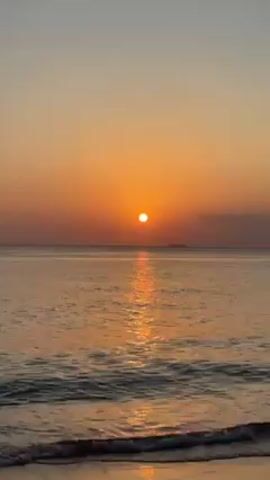 The video shows a sunset over the ocean. The sky is a gradient of orange and yellow, and the sun is a bright orange orb just above the horizon. The ocean water reflects the sunlight, creating a shimmering path of light across the water. Gentle waves are lapping at the shore, and a small dark shape can be seen in the distance.