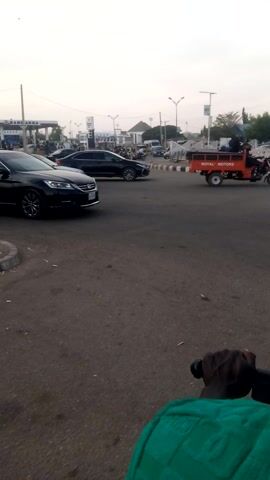 The vertical video captures a street scene from a first-person perspective, likely from a moving bicycle or motorcycle, as indicated by a dark-skinned hand holding black handlebars and a glimpse of a green garment on the right side of the frame. The setting is an asphalt road, appearing dry and well-maintained.

At the beginning of the video, a black car, possibly a sedan or SUV, is seen on the left side of the road, either parked or moving slowly. In the far background, an orange truck with the text "ROYAL MOTORS" partially visible on its side is present. As the camera moves forward, a light blue compact car becomes more prominent on the left, positioned alongside a white and black striped curb or barrier.

A black motorcycle, carrying two dark-skinned individuals dressed in light-colored attire, then enters the frame from the left and crosses the road, moving away to the right. In the distance on the left side of the road, more motorcycles and possibly bicycles can be observed. The background features various buildings, streetlights, and other urban structures. The sky is bright and clear, suggesting it is daytime, possibly morning or late afternoon given the soft quality of the light. The camera continues to move forward throughout the short clip, providing a dynamic view of the street and its traffic.
