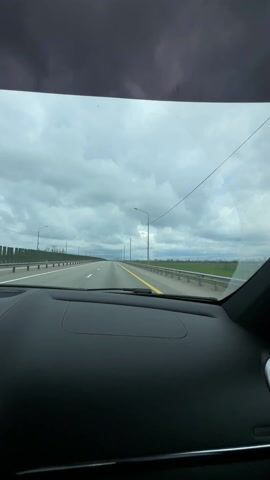 The video shows a view from inside a car driving on a highway. The sky is overcast with dark clouds, but a patch of blue is visible through a gap.  The road is marked with a yellow center line and bordered by a green field on the right.  There are lampposts along the highway, and a metal barrier separates the road from the field. The dashboard of the car is black. Music is playing.
