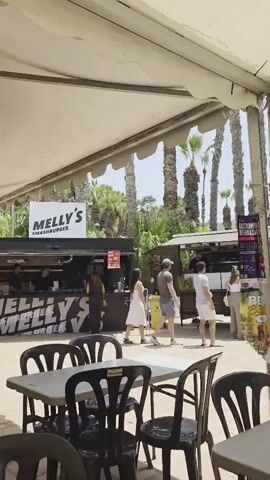 The video shows an outdoor food court with multiple food stalls and seating areas under a large white tent. In the foreground, there are several tables and black plastic chairs. To the left, a man wearing a striped shirt and a red baseball cap sits at a table, facing away from the camera. In the middle ground, there is a food stall labeled "MELLY'S SMASHBURGER" with people queuing and ordering food. Further back, other food stalls are visible, with people interacting and eating. The environment appears to be sunny, with palm trees visible in the background.