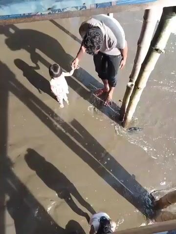 A high-angle, slightly shaky video shows a father and child on a muddy shoreline. The father, wearing a grey t-shirt and black shorts, is holding the hand of a young child dressed in a white outfit. The child is facing away from the camera, towards the water, while the father is facing the child. They are standing near bamboo poles that appear to be part of a structure. The water is shallow and murky, with small waves lapping at the shore. Long shadows stretch across the mud, indicating bright sunlight. In the foreground, another person, a female, is visible from the shoulders up, also appearing to be on the shoreline. The video appears to be a short, handheld recording.