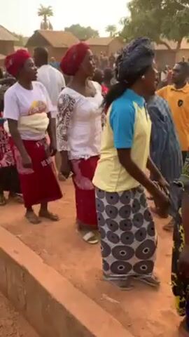 A group of women in a rural area are walking along a dirt road. The women are wearing colorful traditional clothing, including head wraps and long skirts. Some of the women are also wearing white tops. There are other people standing around in the background, and some buildings are visible. The setting appears to be a village or small town in a developing country. The women are moving slowly, possibly in a procession or as part of a cultural event. The atmosphere seems relaxed and communal.