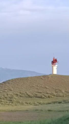 A video displays a gently rolling hill covered in dry grass. A large, light-colored object with a pointed top stands on the crest of the hill. To its right, a tall, arched structure with some rigging is also visible. The sky above is a pale, cloudy blue, and distant mountains can be seen on the horizon. The foreground features a strip of green grass.