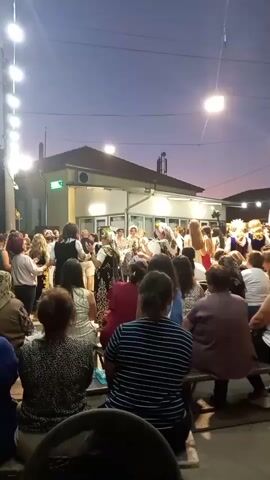 The video captures an outdoor gathering at dusk, illuminated by strings of lights. A group of people are watching performers who are dancing and possibly singing in the center of the scene. The performers are wearing traditional costumes, some with elaborate headwear. The audience is seated and standing, facing the performers. The background features a building with windows and a sign that appears to be green. The sky is a deep blue, indicating the transition from day to night. The overall atmosphere suggests a festive or cultural event.