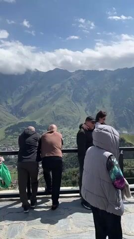 This is a vertical video taken on a sunny day with some clouds, showcasing a scenic mountain landscape. In the foreground, a group of people are standing at a railing, looking out at the view. Two men in the center are looking away from the camera, one wearing a brown jacket and the other a black jacket. To their left, another man in a black jacket and dark pants stands with his back to the camera. To the right, a couple is embracing and looking at a phone, the woman is seated on the railing. In the far right foreground, a person wearing a grey hooded vest with a colorful, galaxy-patterned sleeve is partially visible, facing away from the camera. Further to the right, another person in a green jacket with the word "REPLAY" printed on the back is visible from behind. In the middle ground, a small town is nestled in a green valley, with buildings and roads visible. The background is dominated by majestic, rugged green mountains, with some clouds clinging to their peaks. The camera pans slightly, shifting the focus between the people and the expansive mountain scenery.