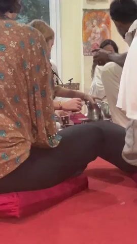 The video shows a person from the knees down, wearing black leggings and a printed shirt, sitting on a red cushion on a red floor. The person's right leg is extended, and their left leg is bent at the knee. In the background, other people are visible, and there are various objects, suggesting an indoor gathering or event. The lighting is dim.