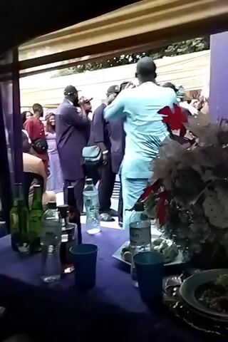 The video shows a group of people standing and talking outdoors, seemingly at an event or gathering. They are dressed in a variety of clothing, including traditional African attire. A table in the foreground is draped with a purple cloth and holds water bottles, alcohol bottles, cups, plates, and a decorative floral arrangement. The atmosphere appears lively, suggesting a social event. The camera is mostly static.