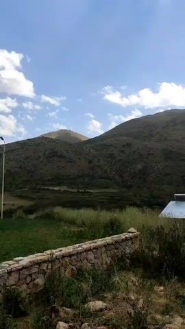 A rural landscape featuring rolling hills under a partly cloudy sky. In the foreground, a low stone wall runs horizontally. To the right of the wall, a small house with a metal roof is visible, along with some vegetation. Further to the right, a white car is parked. The video pans slightly, offering a view of power lines stretching across the frame.