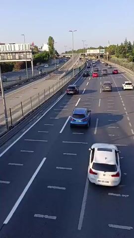 A high-angle, slightly shaky, over-the-shoulder view shows a multi-lane highway with moderate traffic. Cars are traveling in both directions, with vehicles in the foreground and midground moving away from the camera. The road is paved and well-maintained, with clear lane markings. On the left side of the highway, a barrier separates the lanes from what appears to be an access road or another section of highway. Beyond that, there are buildings, trees, and streetlights. The sky is clear and blue. The time stamp indicates the video is from the early morning.
