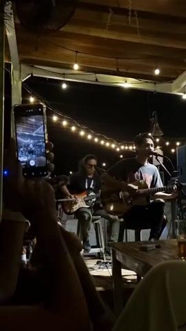 Here's a concise description of the video:

A live band is performing at night under a covered, well-lit outdoor area. Two guitarists are seated on stools in the foreground, one also singing into a microphone. A person in the foreground is recording the performance on a smartphone, the phone's screen showing a live feed of the band. String lights are strung across the ceiling, and a large fan is visible in the upper left corner. Tables and chairs are scattered around the area, suggesting an audience is present. The overall atmosphere appears relaxed and intimate.