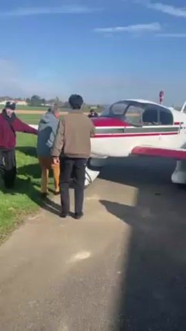 Three men push a small single-engine airplane from a paved surface onto grass. The plane is white with red markings, and the registration number F-BRZQ is visible on the tail. The men are dressed in casual clothing. The sky is blue with some clouds. The background includes a green field and some buildings in the distance. One of the men mentions sending the video to his mother.