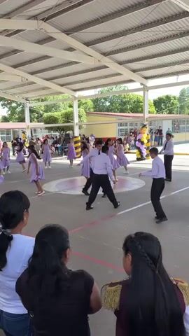 A group of children in light purple dresses are performing a dance on a paved area under a covered roof. Some of the children are in formation, while others are scattered across the area. A few young boys in white shirts and dark pants are also present, some of them appearing to be part of the performance. Several adults are watching the performance from the foreground, with some of them interacting with each other. In the background, there are buildings with yellow and red accents, and trees. The overall environment appears to be an outdoor school or community event.