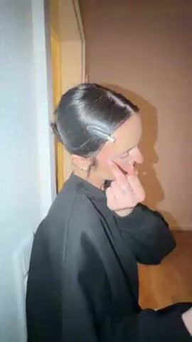 A woman in a black sweatshirt with her hair clipped back shows her middle finger to the camera. The camera pans to a man with curly hair, wearing a vest over a white shirt, looking at his phone. The camera moves in close to his face, focusing on a small red mark on his nose.