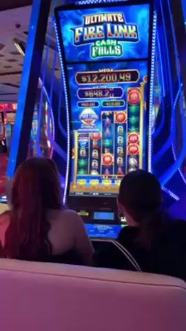 Two women are seated in front of an "Ultimate Fire Link Cash Falls" slot machine in what appears to be a casino. The machine displays digital graphics and jackpot amounts. The screen is illuminated with bright blues and reds. The woman on the left has long reddish-brown hair, and the woman on the right has dark hair. The environment seems to be a bustling casino floor, with other slot machines visible in the background. The women are looking at the screen. The sound of the slot machine is audible.