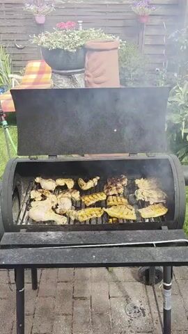The video shows an outdoor barbecue with chicken pieces cooking on a grill. The grill is black and appears to be a smoker-style grill. There is smoke rising from the grill. In the background, there is a wooden fence, some potted plants with pink and white flowers, and a lawn chair with a checkered pattern. The ground is paved with bricks. The scene is lit by natural daylight. The video is shot from a stationary position.