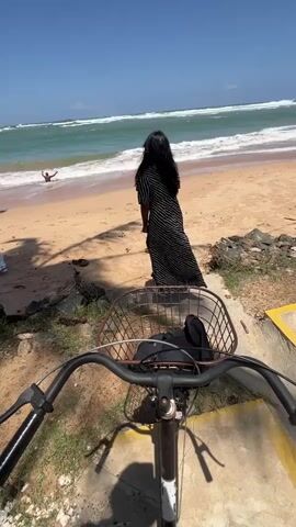 The video shows a first-person perspective of someone on a bicycle facing a beach. In the distance, a woman in a black and white striped dress stands on the sand, facing the ocean. To the left of the woman, in the shallow water, a person is visible in a crouching or sitting position. The ocean has moderate waves. The foreground shows the handlebars and front basket of the bicycle, with some items in the basket. The beach is sandy with some rocks and debris along the edge. The sky is blue and sunny.