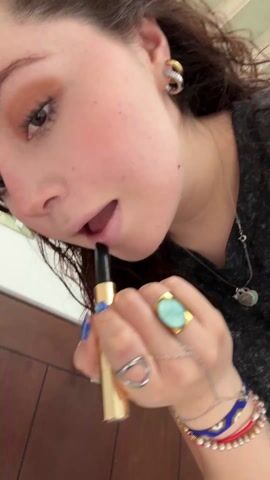 A fair-skinned woman with brown hair and hazel eyes applies a clear lip gloss to her lips. She has a number of rings on her fingers, including a turquoise ring on her right hand. She also wears a silver bracelet with blue beads and a silver necklace with a charm. Her fingernails are painted blue. The background appears to be a bathroom with wood paneling.