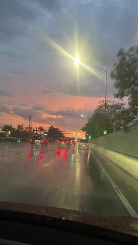 The video shows a view from inside a vehicle driving on a wet road during sunset. The sky is filled with dramatic clouds tinged with orange and pink hues. Traffic lights are visible, some showing green. Several cars are ahead, their taillights reflecting on the wet asphalt. The road is slick with rain, creating reflections of the taillights and traffic lights. Trees and streetlights line the road. The overall mood is atmospheric, with the low light and reflections contributing to a sense of twilight driving.