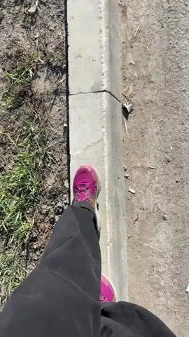 A person wearing dark gray pants and bright magenta athletic shoes walks on a gray concrete curb. The curb is between a grassy area and a paved area. The camera angle is top-down, focused on the person's lower body and the curb. The person walks slowly and deliberately.