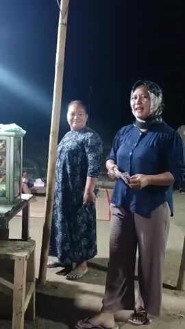 The video opens with a medium shot of two women standing outside at night. The woman on the right is wearing a blue shirt and brown pants, and she is holding some money in her hand. The woman on the left is wearing a dark patterned dress. Behind them, a food stall is visible, with a glass display case filled with food. A bright light illuminates the scene.

The camera then pans to the left, focusing on a younger woman wearing a hijab and a striped shirt, who appears to be serving food from the stall. She is interacting with someone off-camera. The food stall is made of wood and has a clear plastic display case filled with various food items. There are bottles of condiments and a green plastic container on the counter.

The video continues to show the food stall and its surroundings, including a wooden shelf with cooking equipment. A young man in a green t-shirt and shorts is standing to the left of the stall, looking towards the camera. He appears to be a customer. The lighting is dim, indicating it is nighttime. The ground is dirt, and there are various items scattered around, including a woven mat. The stall itself is covered with a light-colored tarp.