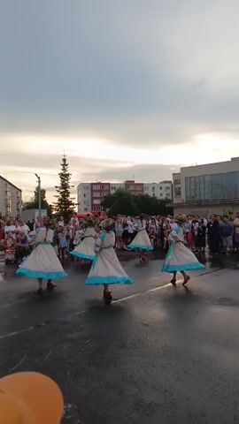 A group of women in traditional folk costumes are dancing in a public square. The costumes are long white dresses with blue trim and red shoes. They are performing a choreographed dance in a circle. A crowd of spectators is watching them from the sides of the square. The square appears to be paved and wet, suggesting it may have recently rained. Buildings, including apartment buildings and a larger modern structure, are visible in the background. The sky is cloudy and overcast, indicating either early morning or late afternoon.