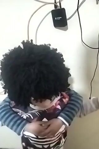 A child with a large afro wig stands with arms folded in front of a white wall. Visible above the child are various electrical cables and a black electronic device. The child wears a long-sleeved shirt with a floral pattern under a blue and white striped shirt. The child's face is mostly obscured by the wig. The camera angle is frontal and slightly above. The scene appears indoors.