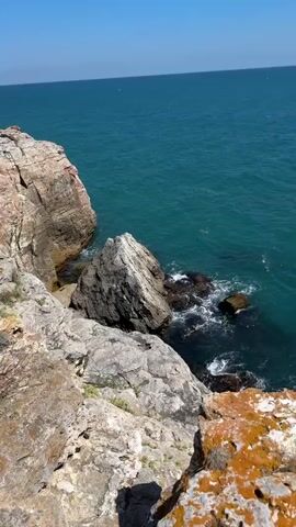 The video shows a panoramic view of a rocky coastline overlooking the ocean. The camera pans from a high vantage point down towards the water, where waves crash against the rocks. The water is a deep turquoise color. In the foreground, there are large, weathered rocks with patches of orange lichen. In the distance, on top of the cliff, there is dry grass and a small building. A single person is visible standing on the cliff edge in the far distance of the frame. The sky is clear and blue.