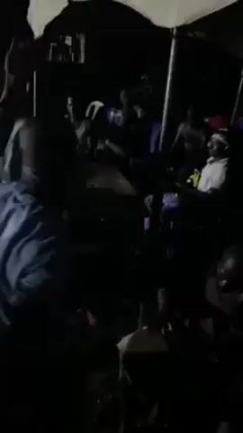 The video shows a dark outdoor scene with a gathering of people seated in chairs under what appear to be tents or canopies. It's difficult to discern much detail due to the low lighting. There are many individuals, but their faces are mostly obscured by shadows. Some people are wearing head coverings. The sounds suggest music playing in the background. Overall, the environment is dimly lit, and the details are hard to make out clearly.