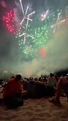 A fireworks display is captured from a crowd of people on a beach. The fireworks are exploding in the night sky, with bursts of red, green, and white light. The crowd is silhouetted against the sky, and some people are sitting and watching the show. There is smoke from the fireworks lingering in the air.