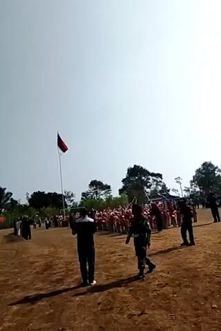 The video captures an outdoor ceremony or event under a bright sky. A red and white flag, likely the Indonesian flag, flies from a tall flagpole on the left side of the frame. In the foreground, two individuals are visible. One, wearing a dark uniform, appears to be holding a camera or recording device. The other, in a dark jacket and pants, stands facing away from the camera. Further back, a crowd of people, many wearing red and white attire, are gathered. Some are standing in formation, while others are observing. Tents or canopies are set up on the left side of the scene, suggesting a festive or organized gathering. The ground is uneven and appears to be dirt or sand. The overall atmosphere suggests a patriotic or celebratory event, possibly a flag-raising ceremony or a performance.
