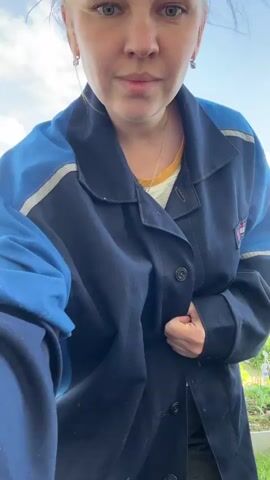 A woman with blonde hair pulled into a bun stands outdoors in front of a blue sky with white clouds. She is wearing a dark blue and light blue jacket with white accents and a yellow shirt underneath. She is holding her arms crossed and looking at the camera. The background shows a garden with rows of plants and some buildings.
