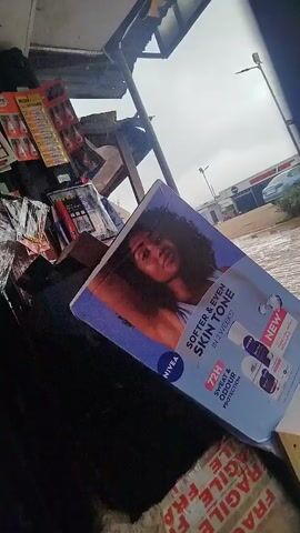The video appears to be filmed from inside a vehicle or structure looking out at a street scene. In the foreground, there is a large advertisement for NIVEA products featuring a woman and several deodorant bottles. The advertisement is positioned at an angle. Beyond the advertisement, the environment appears to be a street with several shops and businesses. There are streetlights visible, and cars are parked or driving on the wet surface of the road, suggesting it may have rained recently. The sky is overcast. There are also some items on shelves in the background to the left, possibly merchandise for sale at a shop. The overall impression is that of a commercial area in an urban or semi-urban setting, possibly in a tropical or subtropical region given the greenery visible.