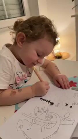 A young child with fair skin and light brown curly hair tied in a bun is seated at a table, focused on tracing a drawing in a coloring book. The child wears a white t-shirt adorned with a pink bow. They are using a light-colored pencil to trace the outline of a chameleon and other figures on the page. The coloring book also has words printed on it. The child has temporary tattoos on their arms. The table is covered with a floral patterned placemat. In the background, a table lamp emits a soft glow, illuminating a small decorative plate with fruit. A window with blinds is also visible. The camera focuses closely on the child’s face and hands as they engage in the coloring activity.