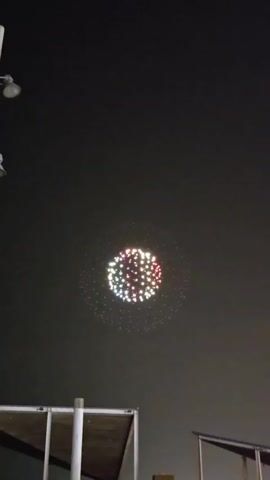The video shows a drone light show at night. A large spherical formation of multicolored lights is visible in the dark sky. The lights are arranged in a dense cluster in the center, surrounded by a sparser ring of lights. The formation appears to be slowly rotating and changing colors. In the foreground, the tops of two structures with white posts and a dark roof are visible, suggesting an outdoor viewing area. The overall impression is of a festive or celebratory event.
