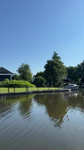 The video opens with a wide shot of a calm body of water, likely a canal or lake, with gentle ripples on the surface. The water reflects the clear blue sky and the surrounding greenery. On the left side of the frame, a modern house with a dark roof and large windows is visible, bordered by a well-maintained lawn and a low fence. Trees and lush vegetation surround the house, creating a serene, suburban or rural landscape. As the video progresses, the perspective shifts, moving along the waterway. We see more of the houses and their manicured gardens lining the water's edge. The houses vary in architectural style, from contemporary to more traditional designs. One house features solar panels on its roof, and a Dutch flag flies from a flagpole in the distance. The final shot reveals the perspective from a watercraft, looking out over the expansive water towards a line of trees and other homes, with the edge of the watercraft visible in the foreground. The overall scene is one of peaceful tranquility on a sunny day, showcasing a picturesque waterside community. There are no visible people in the video, and no overt actions or interactions are captured.