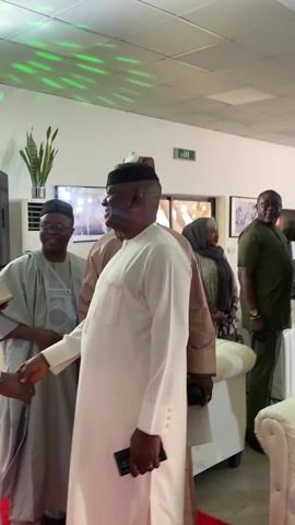A middle-aged Black man in a white robe and black cap, identified as a VIP based on the presence of a bodyguard in camouflage, is walking through a room. He shakes hands with people as he passes. The room is crowded with attendees who are dressed in a mix of traditional and modern attire. The room is well-lit, with a large sound speaker, potted plant, wall art, and other miscellaneous furniture. There is also an "exit" sign.  The VIP smiles and appears to be heading to a formal event.