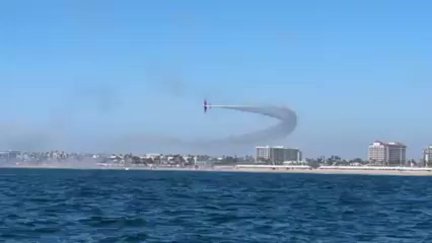 The video shows an airplane performing an aerial maneuver over a body of water, likely an ocean, with a coastline visible in the background. The plane, which appears to be red and white, leaves a trail of white smoke as it flies. The coastline is populated with buildings and palm trees, suggesting a coastal city. The sky is clear and blue. The water is a deep blue, with gentle waves. The camera seems to be positioned on a boat or another vessel on the water.