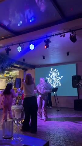 A group of people are dancing on a dance floor in a dimly lit room. The room is decorated with colorful lights and a large screen displays a graphic. The people are wearing casual clothing and appear to be enjoying themselves. Some people are dancing alone, while others are dancing in pairs. The atmosphere is festive and energetic.