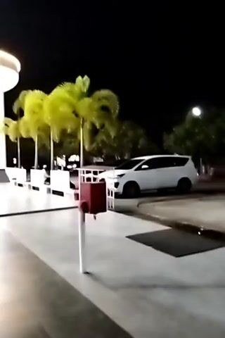 The video shows a nighttime view of a building's exterior. The building has modern architecture with white columns and archways. The area outside the building is well-lit, with lights illuminating the facade and the surrounding paved area. Several palm trees are planted along the building's edge. A white van is parked in the background. The ground is wet, reflecting the lights. A red trash bin on a pole stands in the foreground. The camera pans slightly to the left, showing more of the building's entrance.