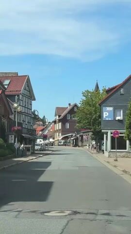 The video shows a street scene in a town with traditional architecture. The street is paved and appears to be a main thoroughfare. On the left side of the street, there are buildings with half-timbered facades and red-tiled roofs. Some of these buildings appear to be shops or cafes, with awnings and outdoor seating. There are people walking on the sidewalk and standing in front of the shops. A silver car is parked on the side of the road. On the right side of the street, there are more buildings with dark siding and red-tiled roofs. One building has a parking sign with a "P" symbol and "90m" indicating a parking area 90 meters away. There are trees and greenery visible in the background, suggesting a hilly or mountainous terrain. The sky is clear and blue, indicating a sunny day. The overall impression is a quaint European town on a pleasant day.