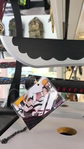 The video shows a display rack of toy anime-themed katana swords in a shop. The swords are mounted on a black rack labeled with Asian characters. The sword in the foreground has a black handle with a red diamond design and a gold cross guard. A tag with anime characters hangs from the handle. A red sword with black detailing and another hanging anime character tag are placed above the black sword. Tactical backpacks are shown on the rack behind the swords. Other swords are visible out of focus in the background. The video is taken from eye level.