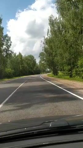 The video shows a view from the driver's seat of a car moving along a paved road. The road is flanked by trees on both sides, creating a tunnel-like effect. The sky is blue with fluffy white clouds. The road has white lane markings. The time stamp indicates the video is playing in real-time.
