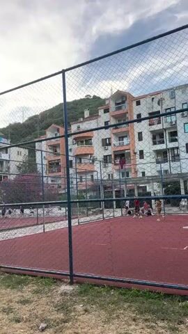 A group of young people are playing basketball on an outdoor court. The court has a red surface and is enclosed by a chain-link fence. In the background, there are apartment buildings and a hill covered in trees. The sky is cloudy. One person is dribbling a basketball, while others are positioned on the court, possibly waiting for the ball or preparing to play. The atmosphere appears casual and recreational.