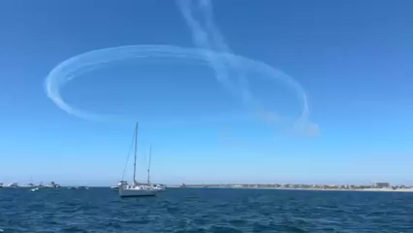 The video shows a daytime scene of a body of water, possibly an ocean or bay, with a clear blue sky above. Several sailboats are visible on the water. In the sky, there is a large, circular contrail, indicating the path of an aircraft performing an aerial maneuver. The coastline with buildings and trees is visible in the background.