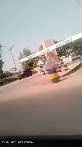The video shows a street scene from the perspective of a moving vehicle. In the initial frame, we see a white archway with "NIGERIA POLICE DUTSEKANO" inscribed on it. A dark-colored truck is parked under the archway. As the vehicle moves, the view shifts to the right, capturing a roadside scene. We observe a low white wall, possibly part of a perimeter fence, running along the roadside. Behind the wall, there are green trees, suggesting a wooded area or possibly a park. The road is paved and appears to be in reasonably good condition. Scattered along the roadside are cylindrical objects with yellow, blue, and green paint, possibly waste bins. The sky is a clear, pale blue. The camera angle is slightly tilted and shaky, indicating the movement of the vehicle. The video's timestamp is at the bottom left corner, indicating "00:00", and below that, a watermark reads "SHOT ON PES-".