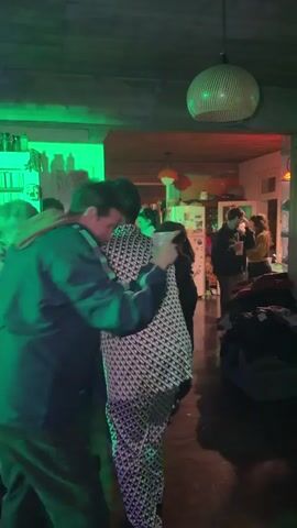 People are dancing at a party in a house, illuminated by multicolored lights. The dancers are of diverse ages and wearing varied clothing. Some people are standing around talking and drinking. The furniture is normal household items.